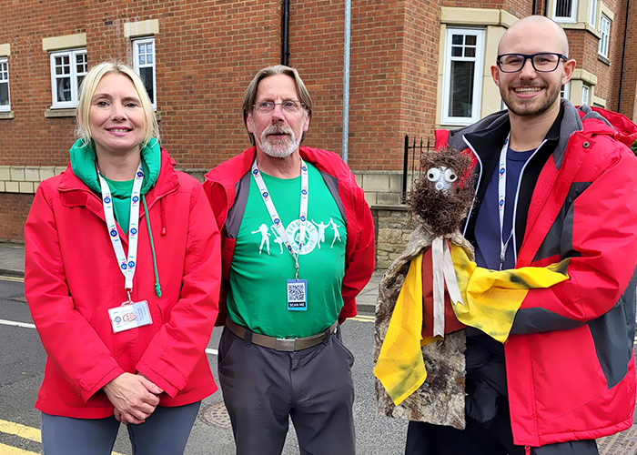 Two white males, one is holding a puppet, and a white female in red jackets and green branded t-shirts with Barnabus safe and sound printed on them