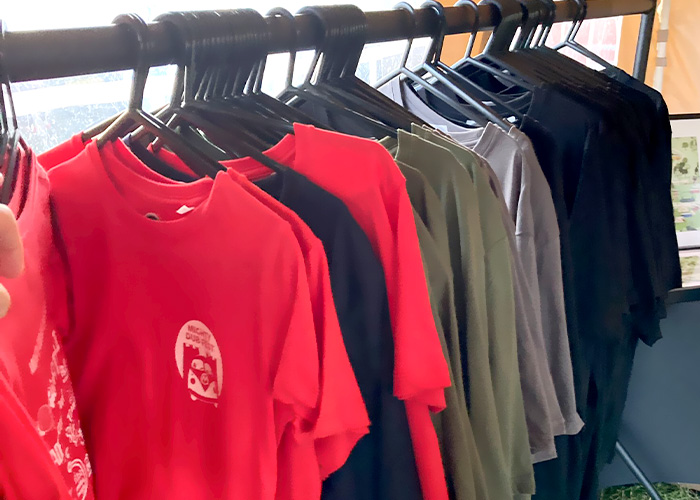 A selection of a dozen or so t-shirts in red, black, brown and purple all with Mighty Dub fest branded on them, hanging on a black clothing rail