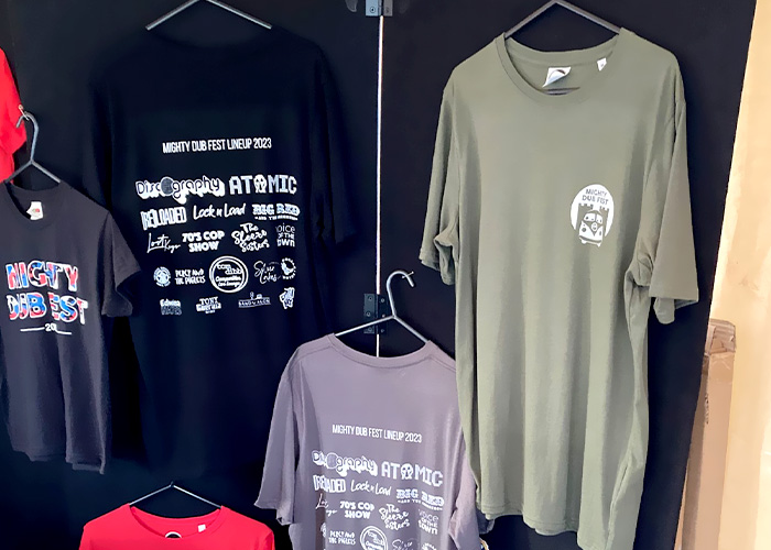 A selection of Mighty Dub Fest branded t-shirts, in purple, navy, black, grey and red, hanging displayed on a black background with clothes hangers